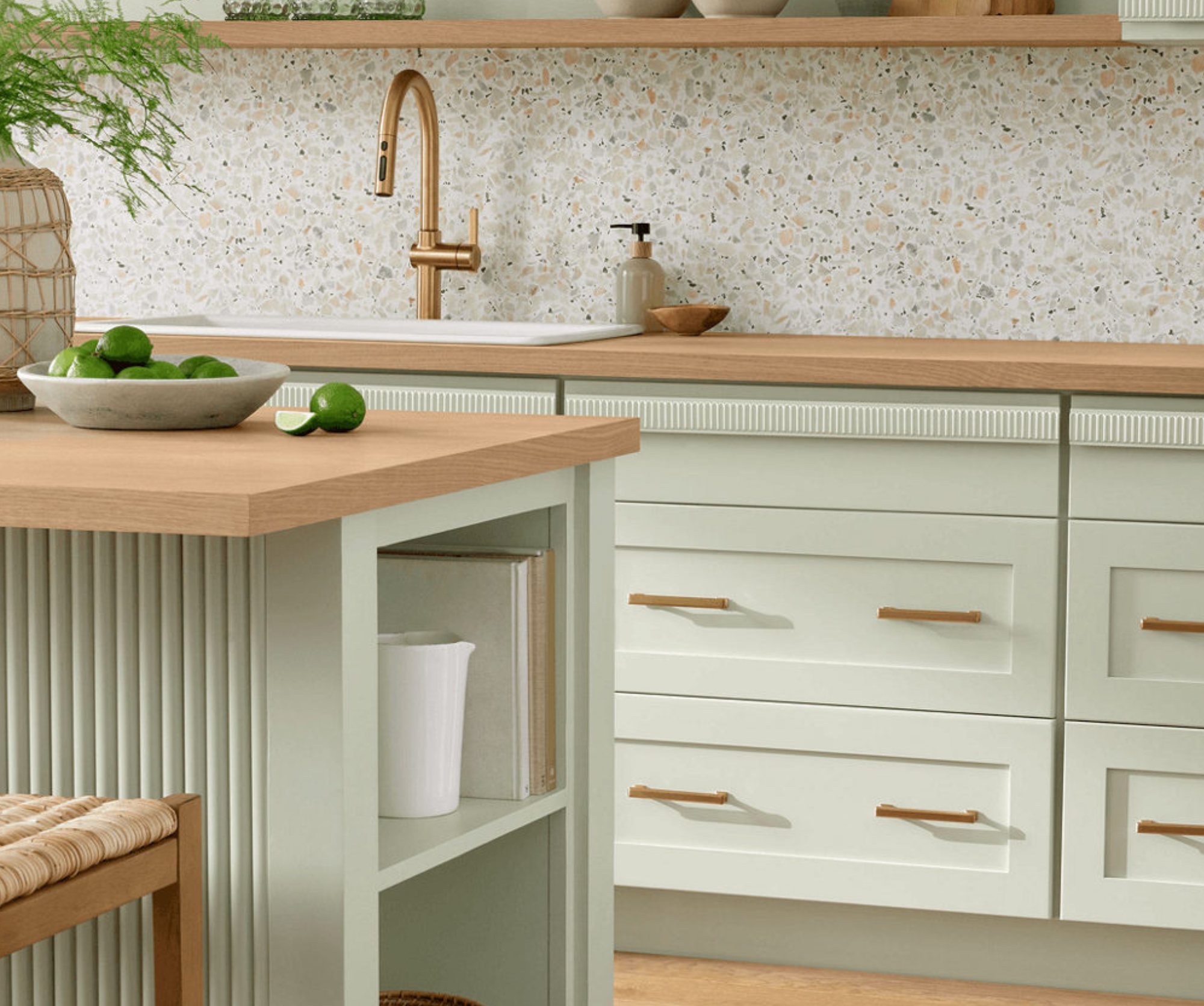 A close-up of a sage green kitchen painted in Liveable Green, with light oak wood countertops, a terrazzo backsplash, a brushed gold faucet, and a bowl of limes on the island.