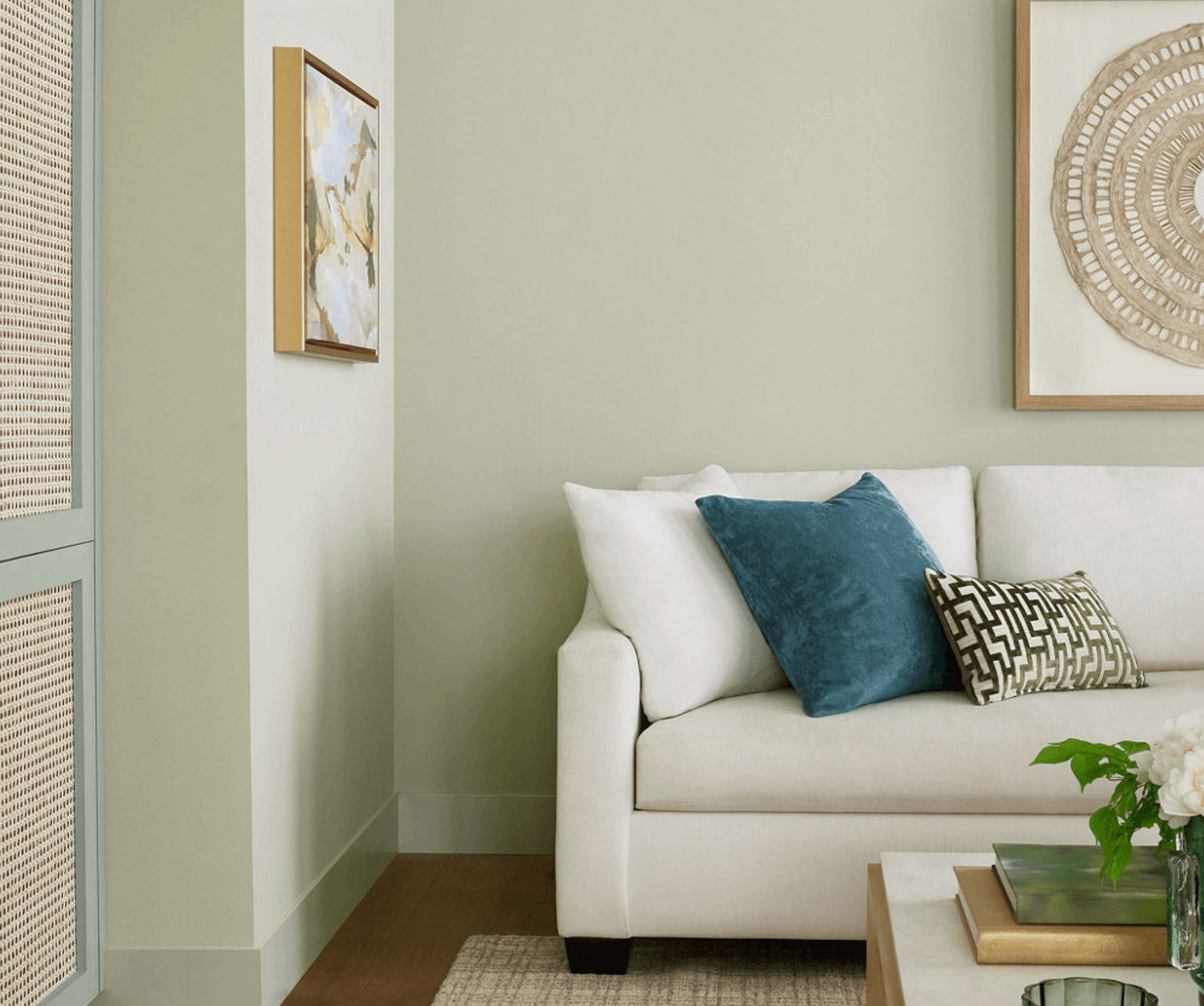 A bright living room corner painted in Liveable Green, with a white sofa, a teal accent pillow, framed artwork, and a wood coffee table with fresh flowers.